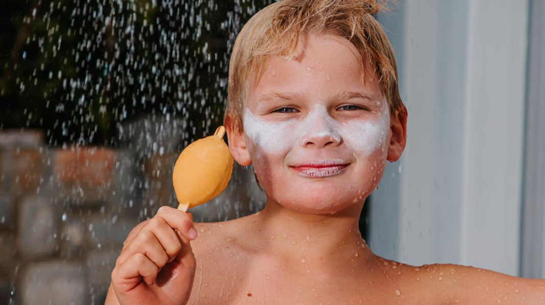 A Boy under a shower wearing white sunscreen on his face while eating an ice cream