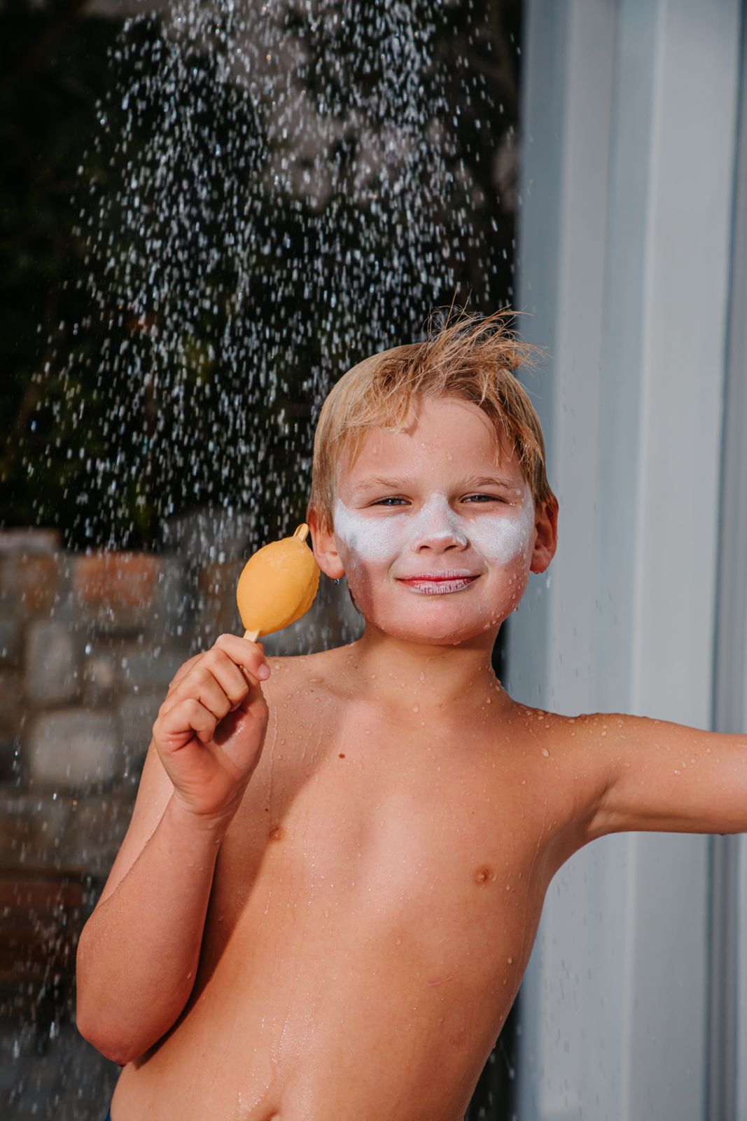 A Boy under a shower wearing white sunscreen on his face while eating an ice cream 